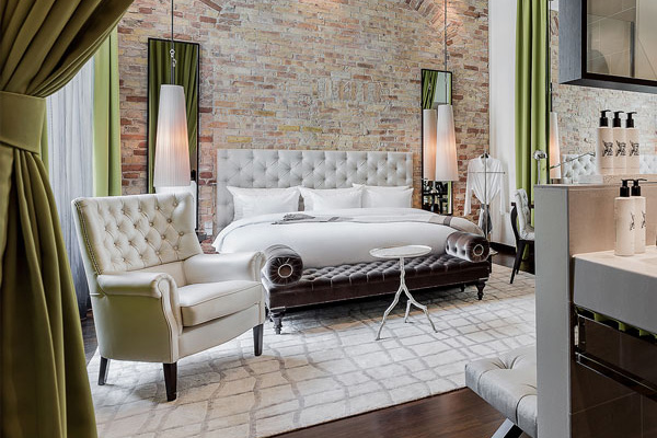 Einblick in die helle Junior Suite mit großzügigem Bett an einer Backsteinwand zwischen zwei Spiegeln und einem weißen gesteppten Sessel